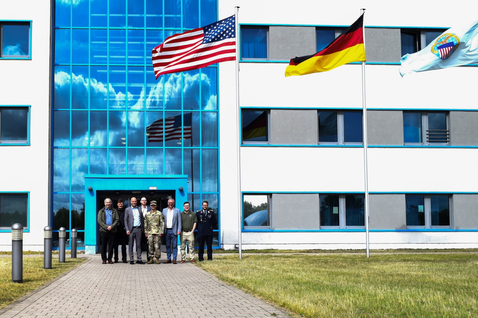 A group of people stand in front of a large building on a sunny day with the American, German and Defense Logistics Agency flags flying in front of the building.