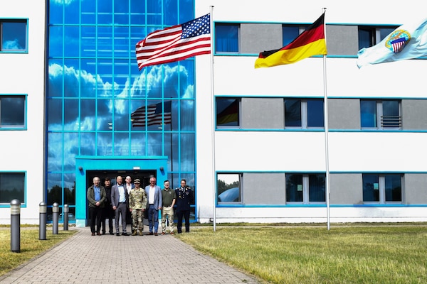 A group of people stand in front of a large building on a sunny day with the American, German and Defense Logistics Agency flags flying in front of the building.