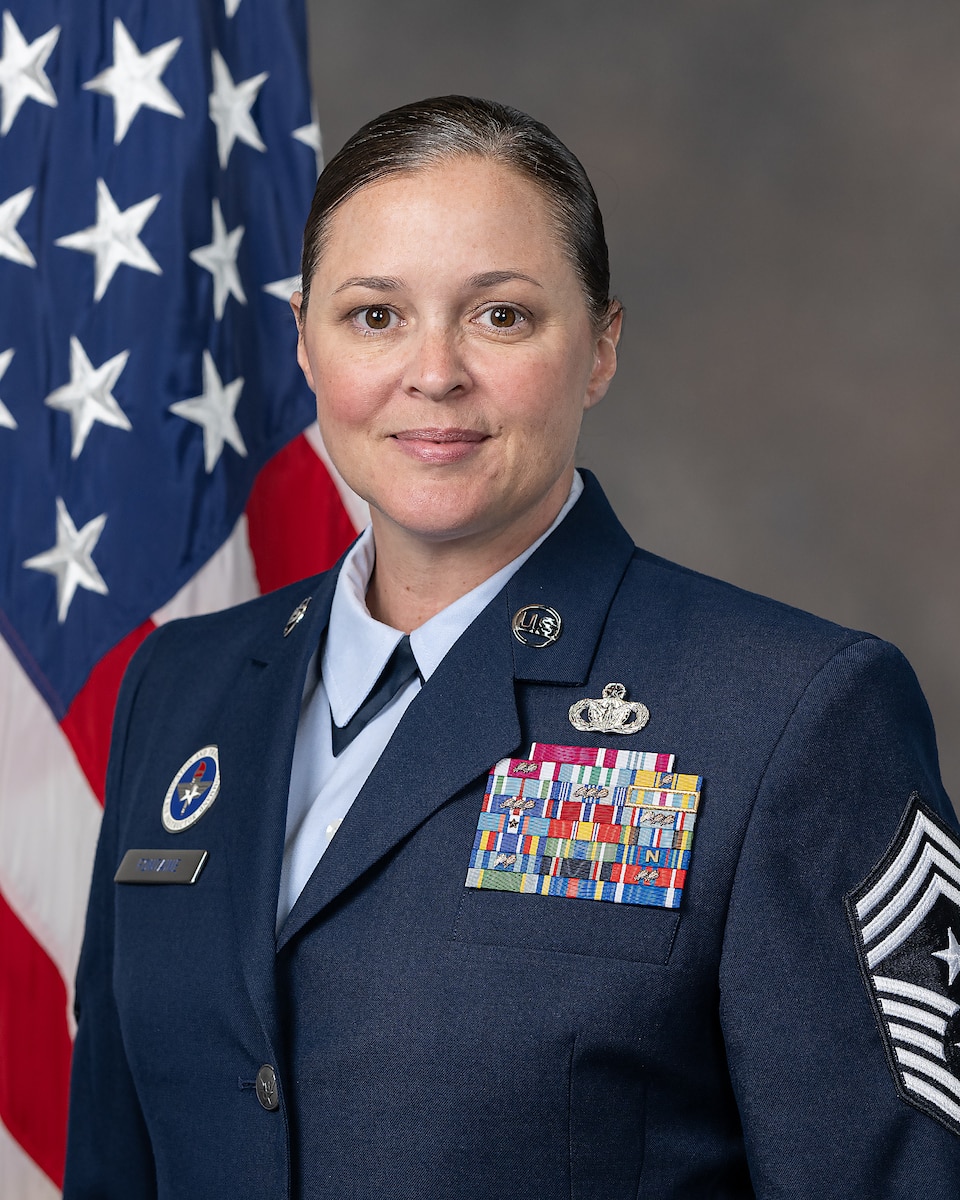 Woman in military dress, standing in front of a gray background with the United States flag to her right.