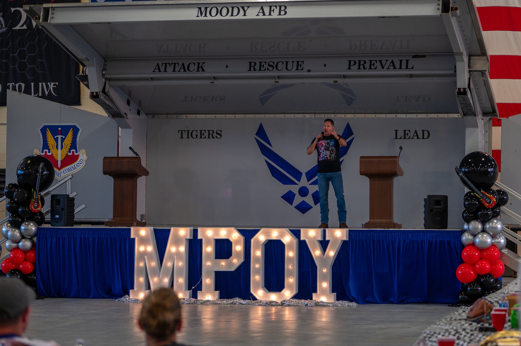 An elevated stage holds a man speaking to a crowd . Big illuminated letters M, P, O and Y, stand in front of the stage.