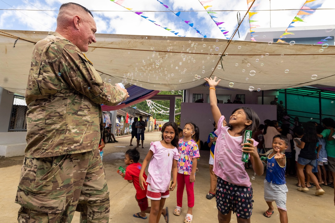 A soldier squirts a bubble maker toward a group of smiling children as they try to catch the bubbles outdoors.