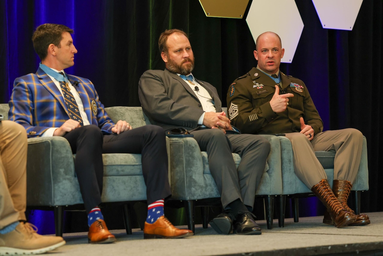 Three men, two in civilian business attire and another in dress military uniform, are sitting next to each other in chairs. One of the men is gesturing with his hands and talking.