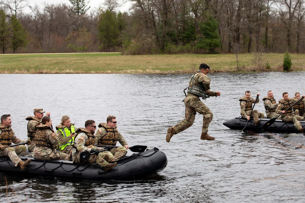 Soldiers using oars in inflatable boats watch as a fellow soldier jumps into the water on a gloomy day with a wooded area in the background.