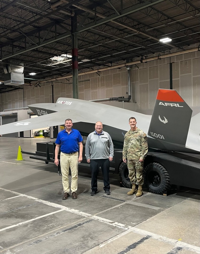 Three men stand in front of the XQ-58 Valkyrie