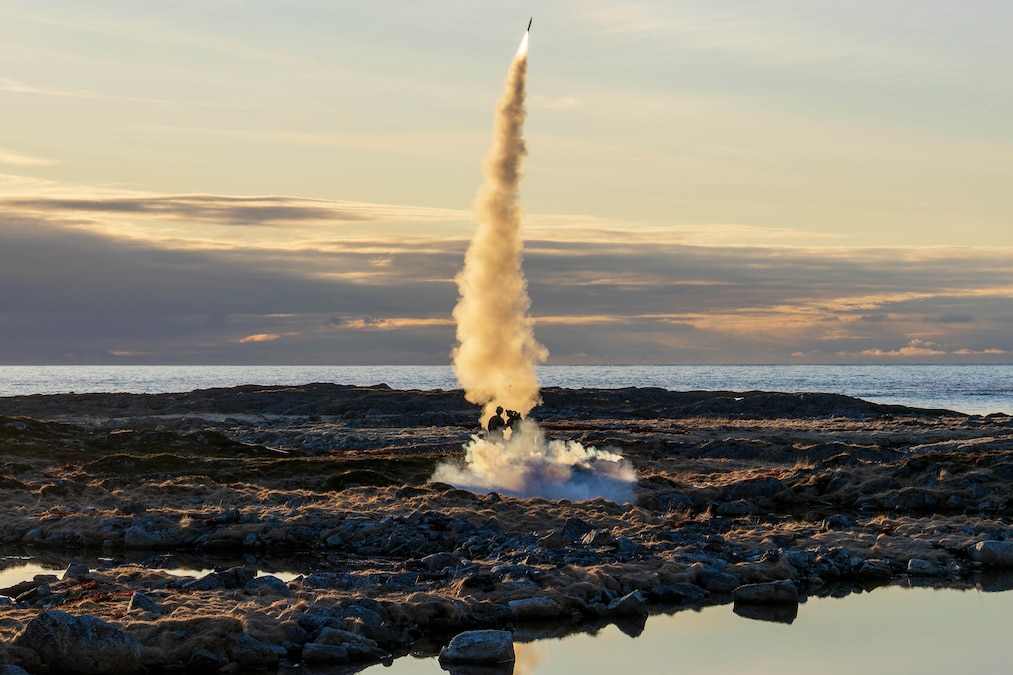 Marines fire a missile from flat, rocky terrain surrounded by bodies of water with a cloudy sky in the background.