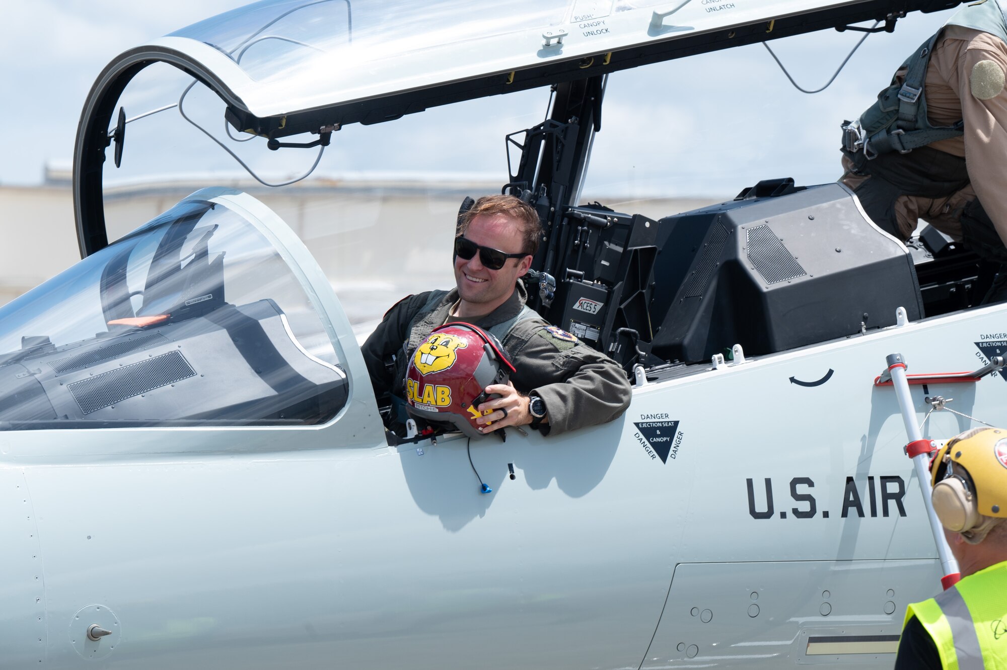 A pilot sits in the cockpit, holding his helmet and smiling.