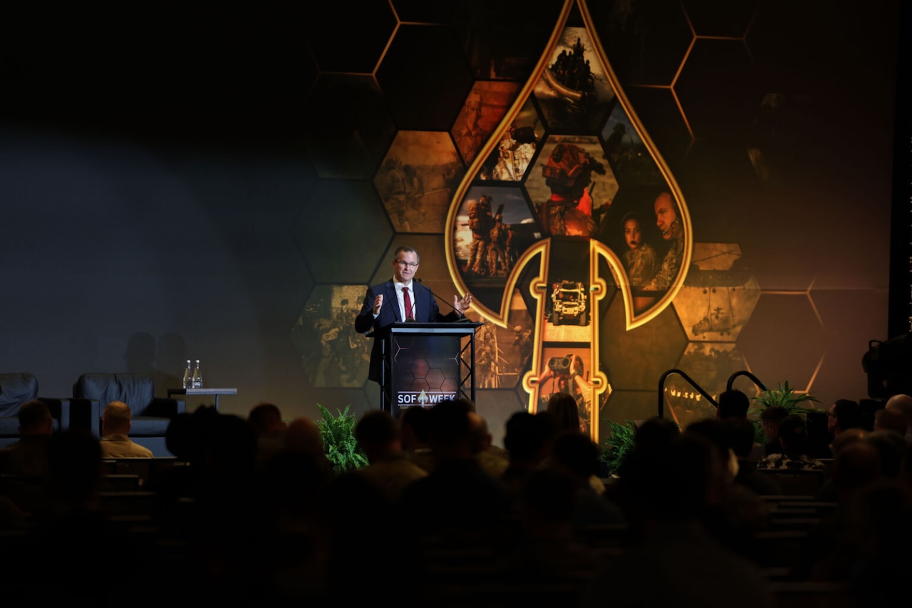 A man in a business suit is standing at a podium on a stage addressing a crowd of people with their backs to the camera. There is a sculpture of a large spear tip next to the man.