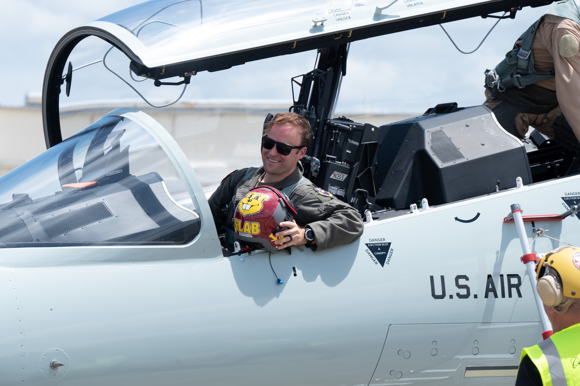 A pilot sits in the cockpit, holding his helmet and smiling.