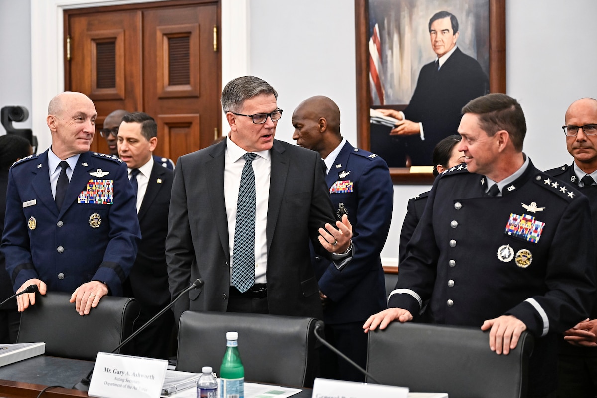 Three Air Force leaders, two in uniform, speak to each other in a hearing room as others stand behind them.