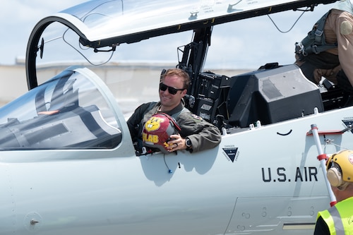 A pilot sits in the cockpit, holding his helmet and smiling.