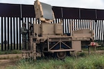 A soldier stands on military equipment outside next to a fence.