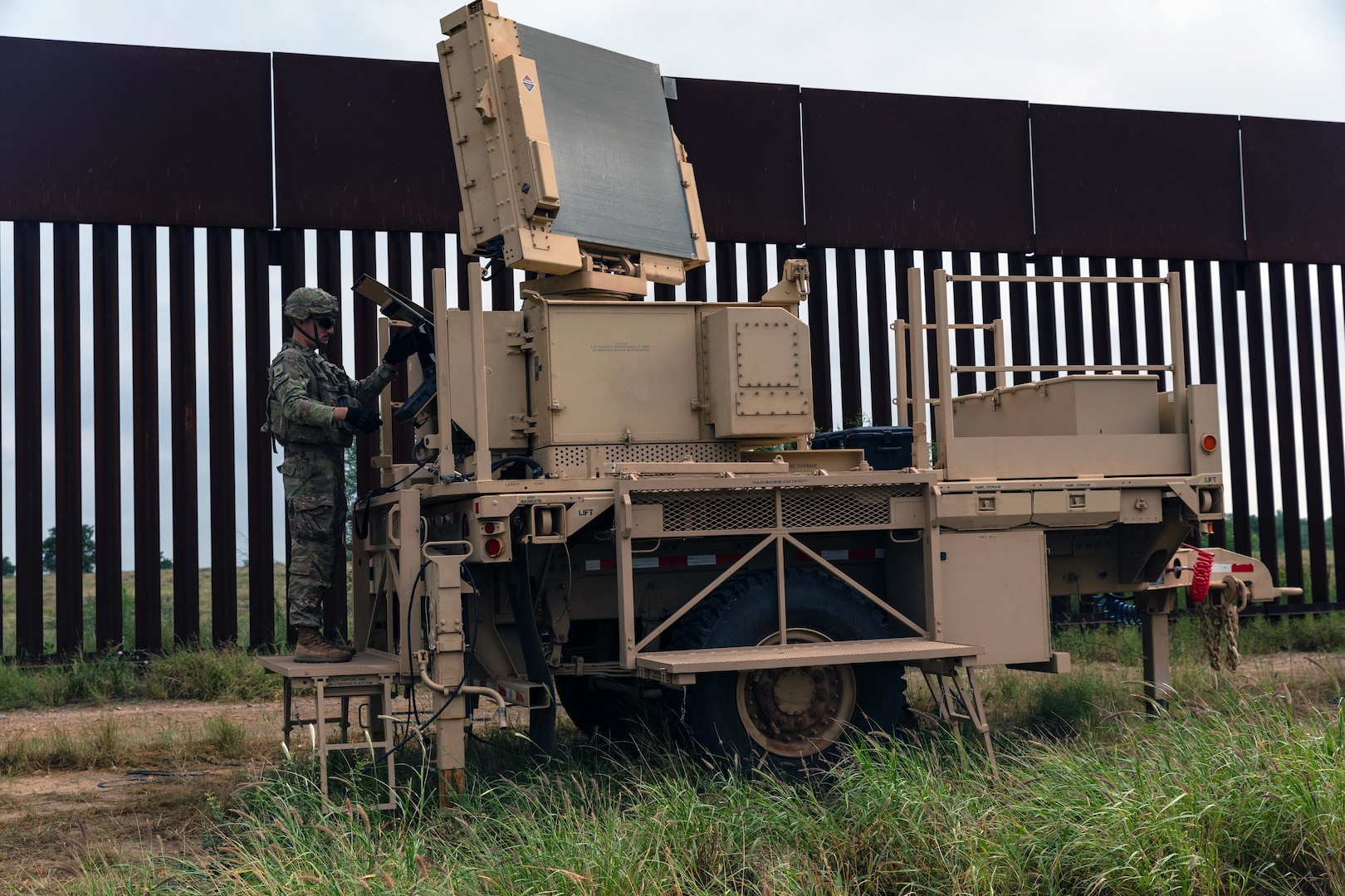 A soldier stands on military equipment outside next to a fence.
