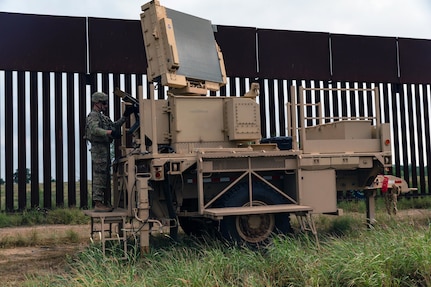 A soldier stands on military equipment outside next to a fence.