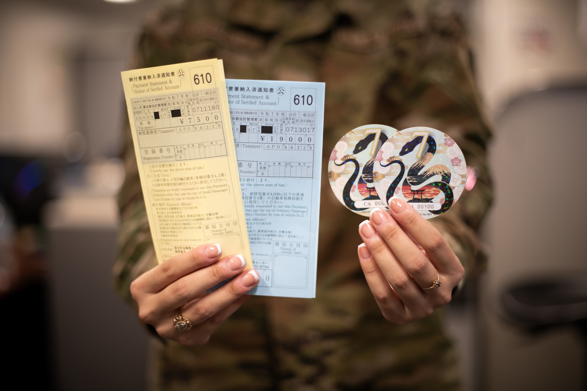 An Airman holds out yellow and blue documents in one hand and the 2025 Road Tax Rodeo window decal in the other.