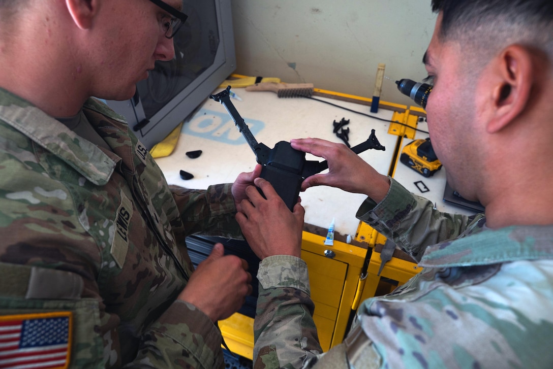 Two soldiers work on a drone above a workbench.