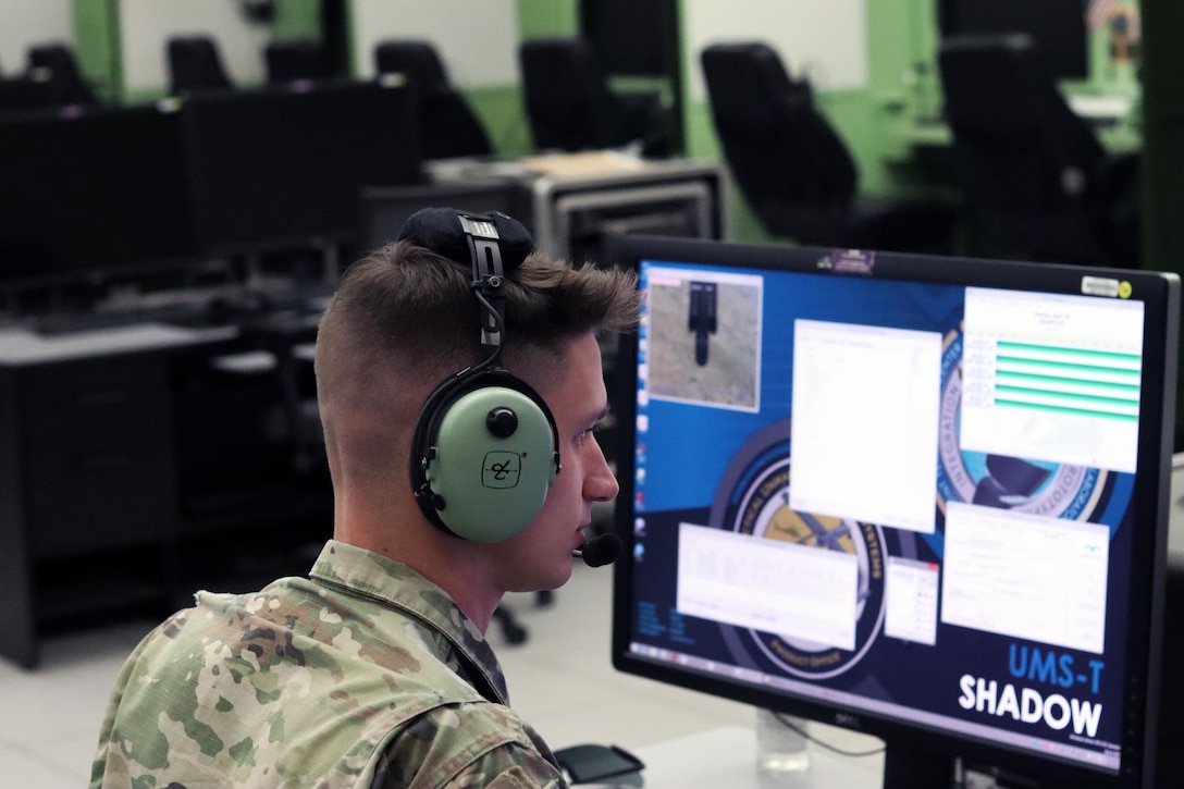 A soldier wearing a headset sits in front of a computer monitor.