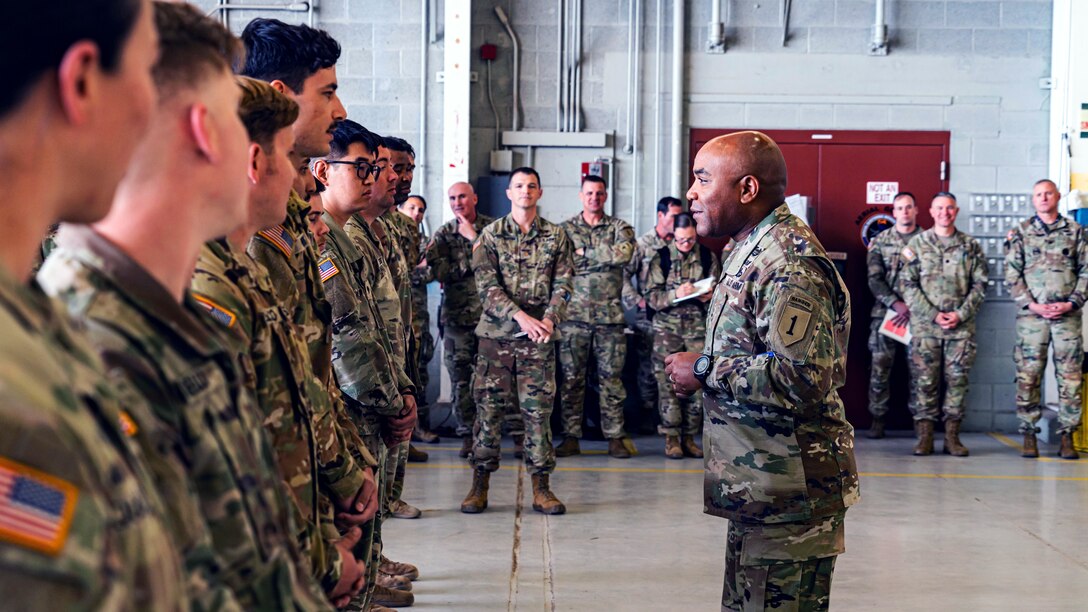 A general speaks to soldiers standing in a line as other soldiers stand in the background.