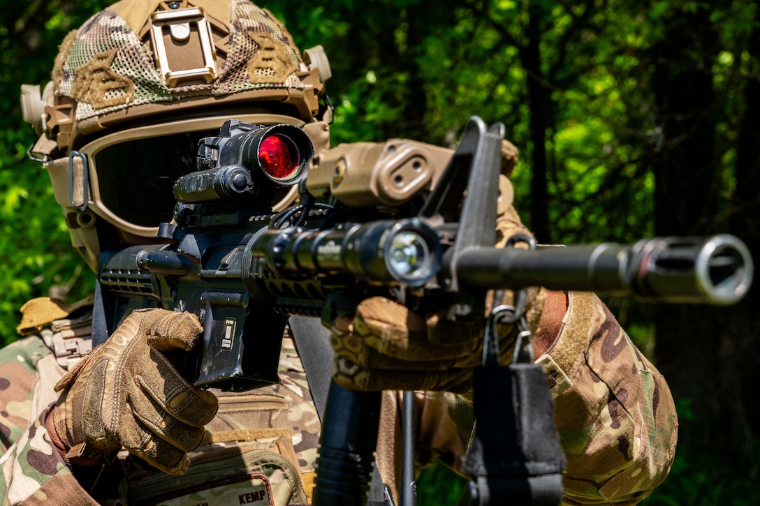 A close-up of an airman in tactical gear looking through the red scope of a weapon with a wooded area in the background.