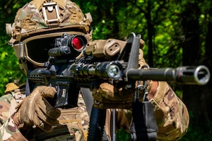 A close-up of an airman in tactical gear looking through the red scope of a weapon with a wooded area in the background.