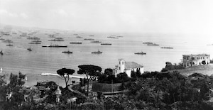Overlooking the hill with several houses and a church are dozens of ships that fill a harbor.