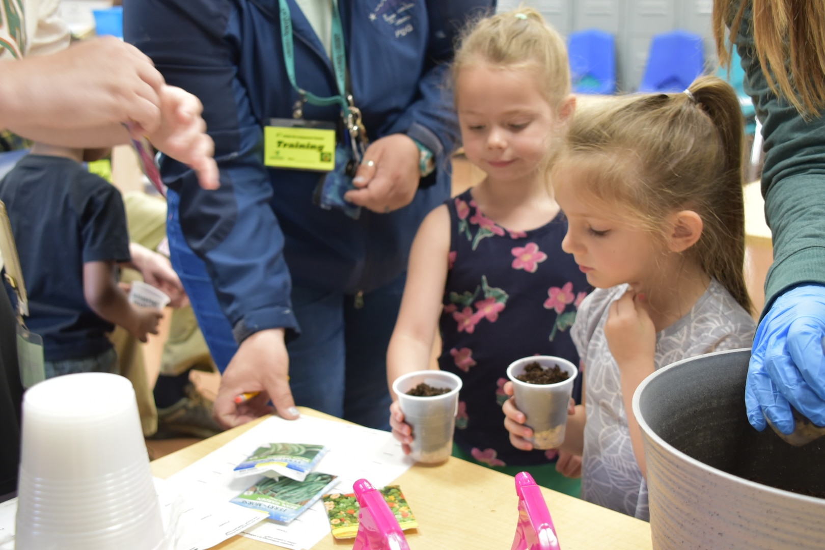 Two children hold cups of dirt while looking at seed packets on a table.