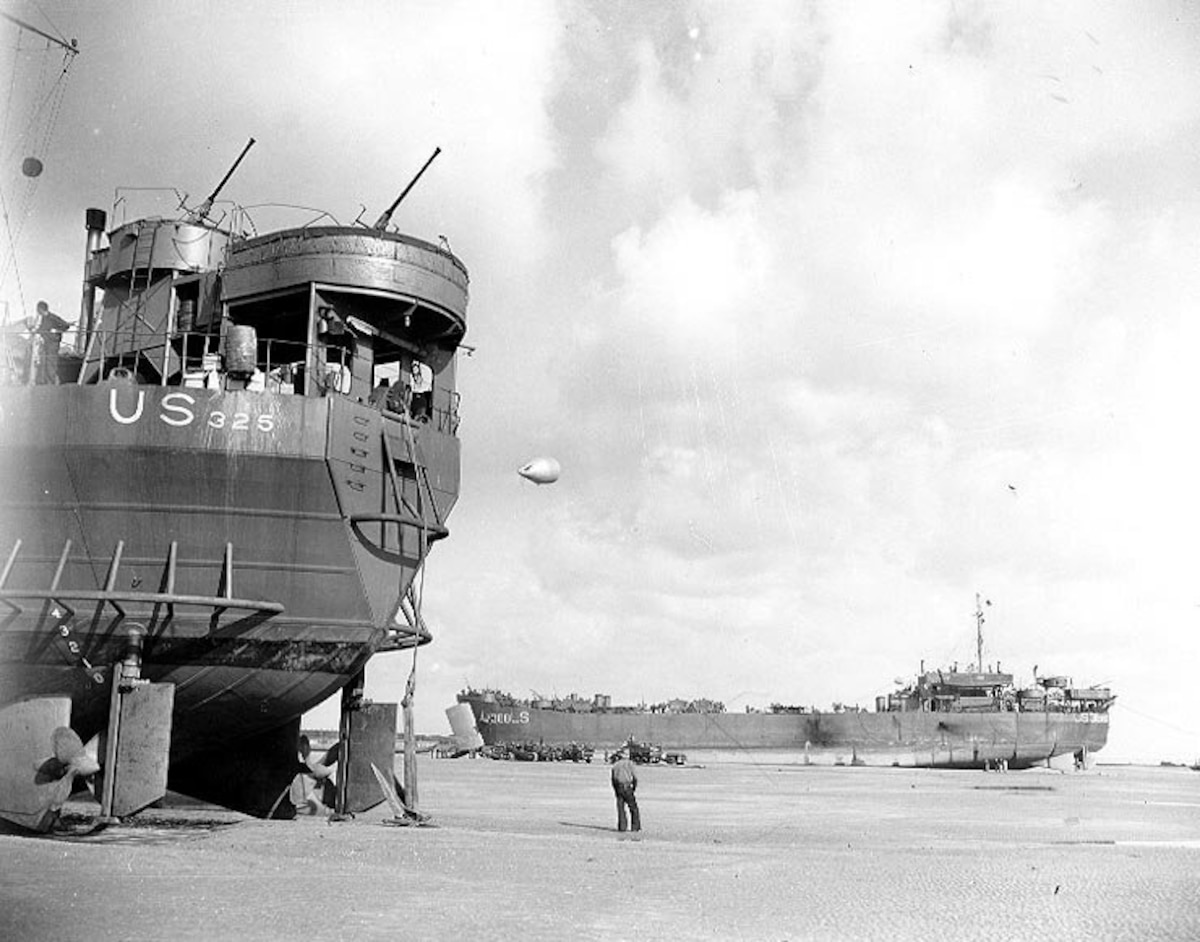 A man stands in shallow water in between two large ships that are pulled up on a beach. There is a weather balloon in the air.