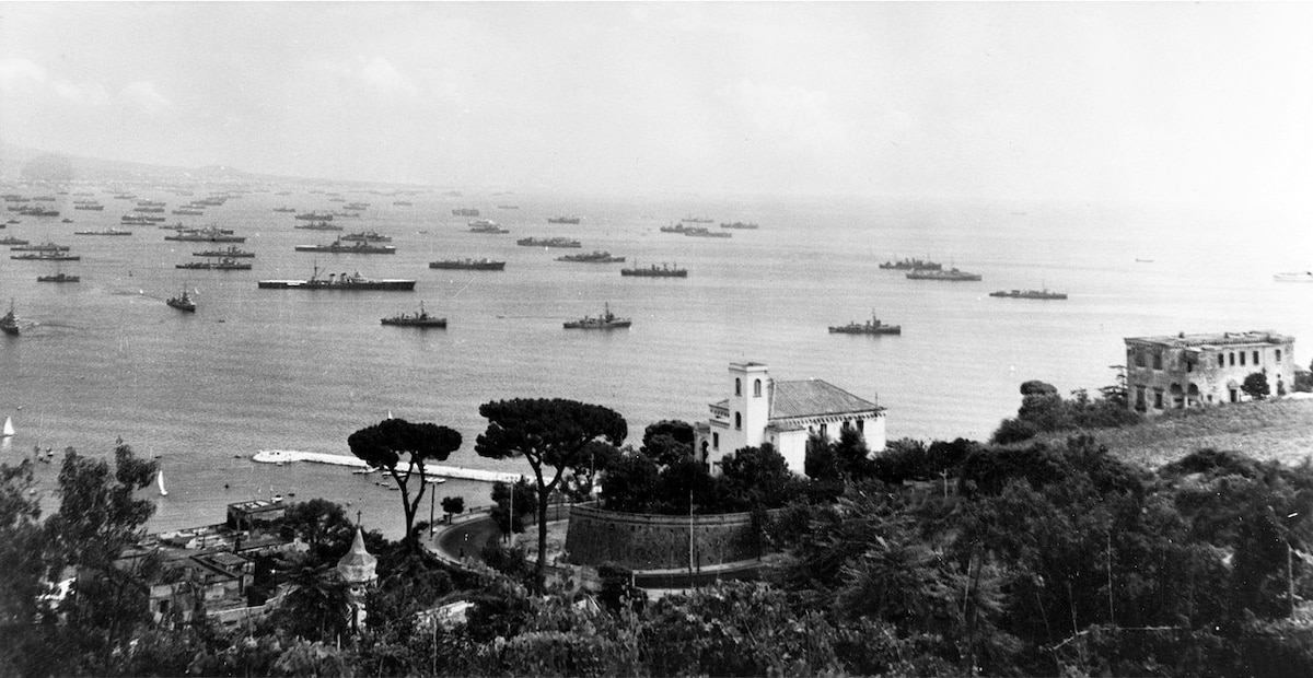 Overlooking the hill with several houses and a church are dozens of ships that fill a harbor.