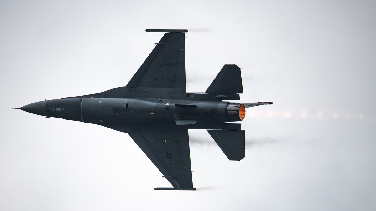 an F-16 fighter jet flies across the sky, afterburner flames visible from the engine nozzle