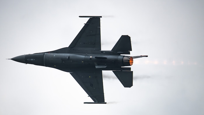 an F-16 fighter jet flies across the sky, afterburner flames visible from the engine nozzle