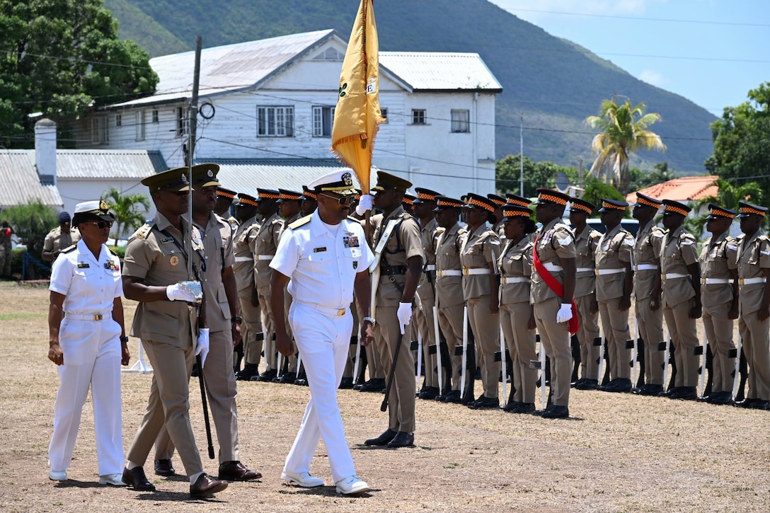 A man in a while military uniform walks and reviews a formation of service members in brown uniforms.