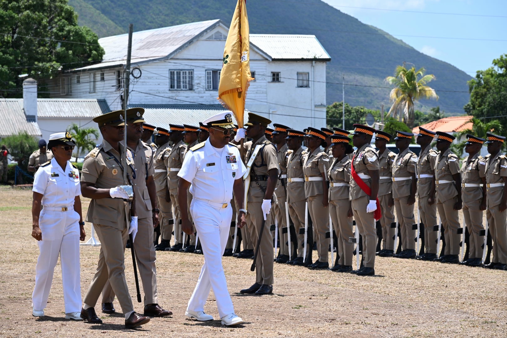 A man in a while military uniform walks and reviews a formation of service members in brown uniforms.