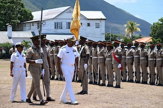A man in a while military uniform walks and reviews a formation of service members in brown uniforms.