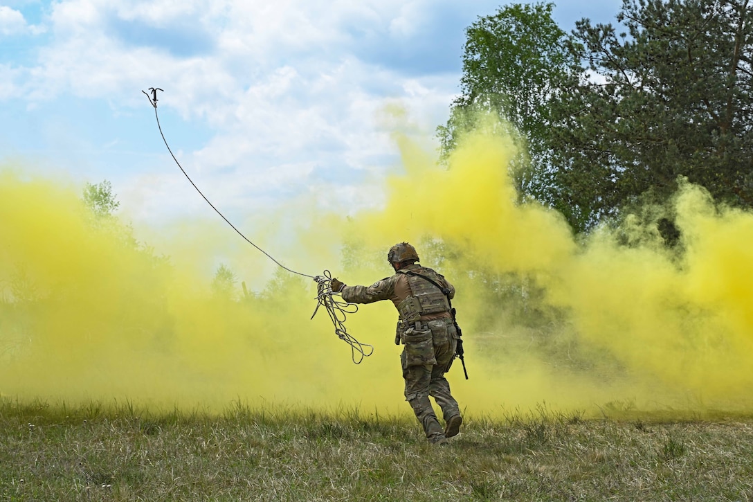 A soldier throws a line into yellow clouds of smoke while standing in a field under a cloudy, blue sky.