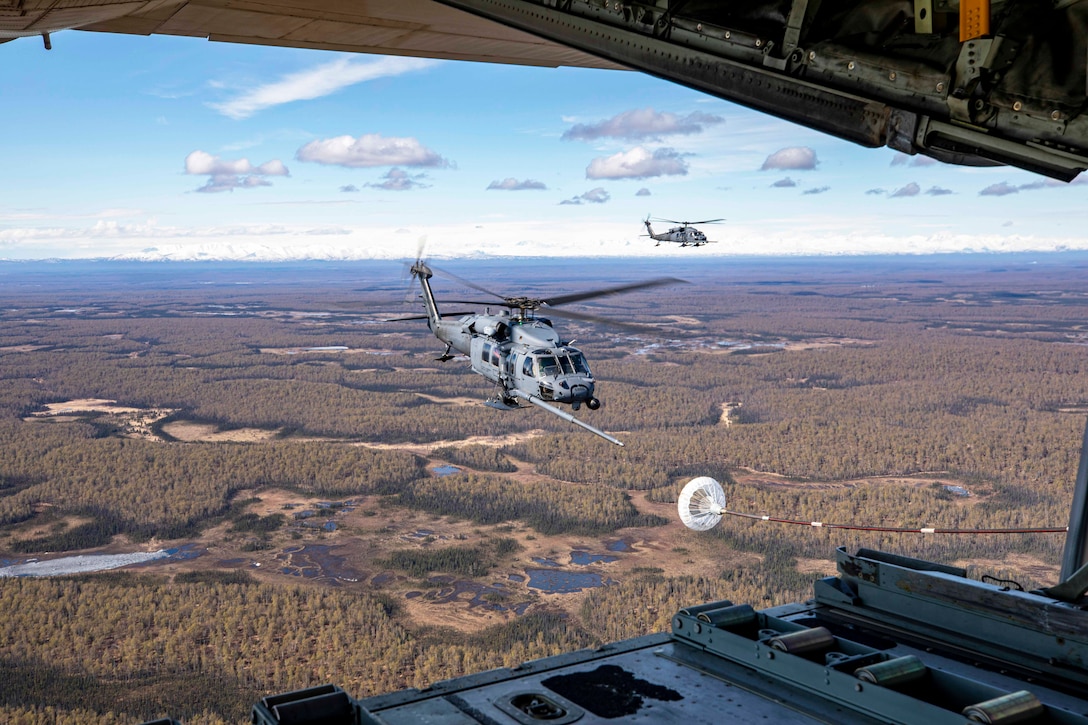 An aircraft with an open rear door extends its boom to another aircraft over a wooded area as another aircraft flies in the distance against a blue, cloudy sky.