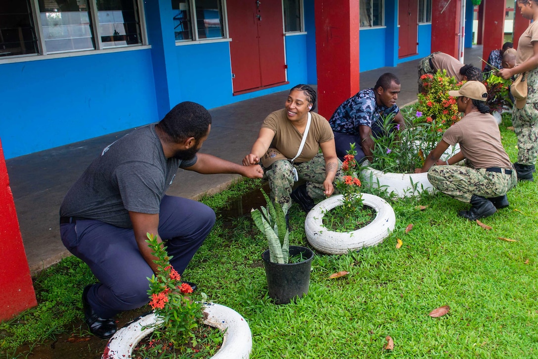 U.S. and Fijian sailors squat while planting colorful flowers outside of a blue and red building.