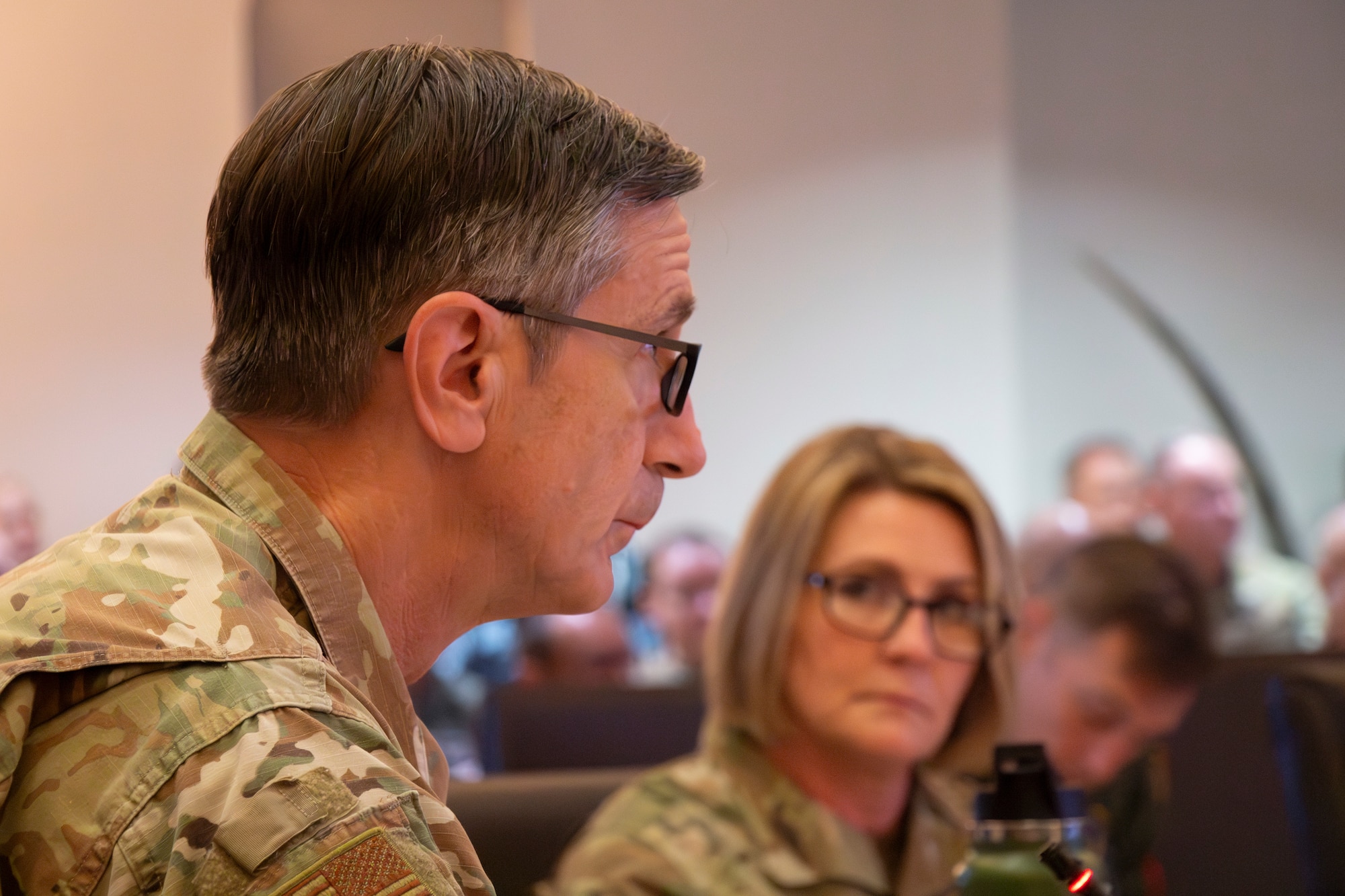 A bespeckled older man in uniform looks up at a screen while a woman in glasses loos at him from across the room.
