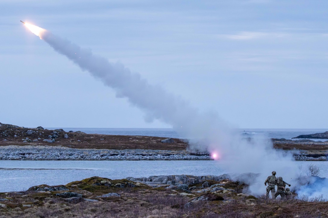 Soldiers fire a missile, from rocky terrain near a body of water, leaving a trail of smoke as it ascends.
