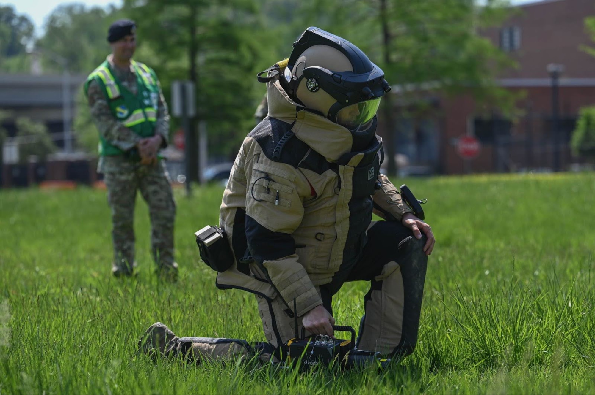 Airman wearing EOD suit kneels facing right as an inspector wearing a green vest observes.