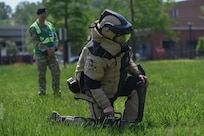 Airman wearing EOD suit kneels facing right as an inspector wearing a green vest observes.