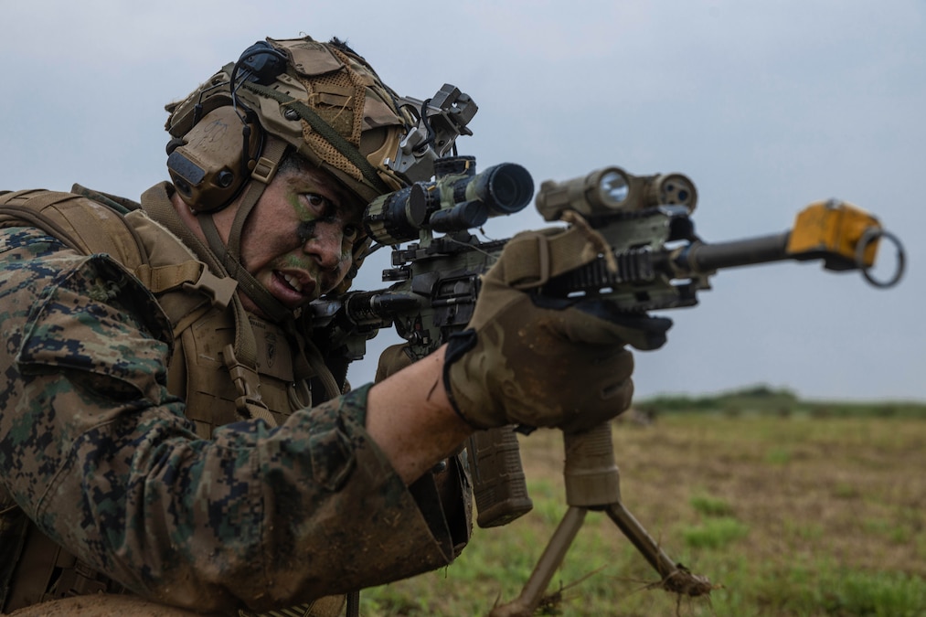 A Marine wearing combat gear aims an automatic rifle from grassy terrain on a cloudy day.