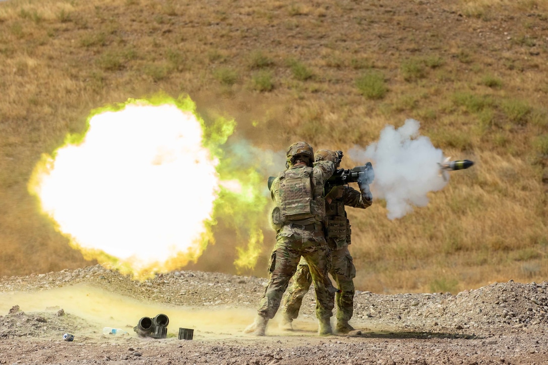 Two soldiers stand next to each other and fire a weapon in the desert during the day causing a large fiery explosion and smoke.