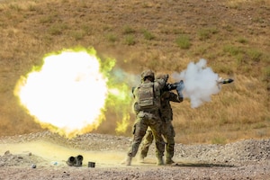 Two soldiers stand next to each other and fire a weapon in the desert during the day causing a large fiery explosion and smoke.