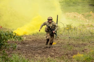 A soldier in combat gear sprints forward in an open field with green foliage and thick yellow smoke rising behind.