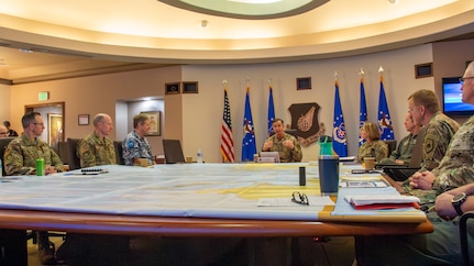 Eight people in uniform sit around a large, square table with a map covering the surface.