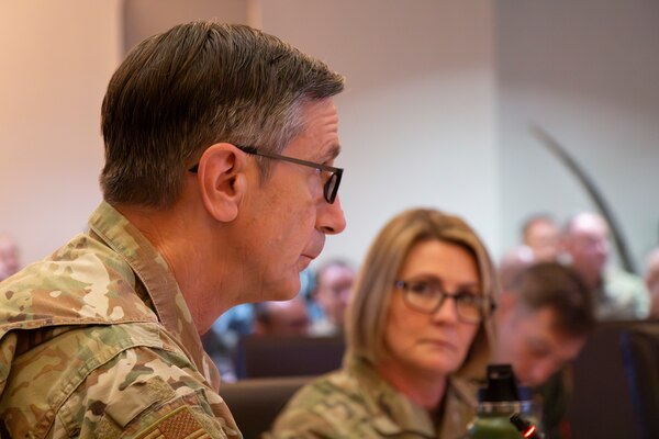 A bespeckled older man in uniform looks up at a screen while a woman in glasses loos at him from across the room.