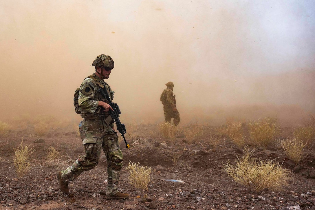 Dust blows in the air as two soldiers in tactical gear walk through a desert.