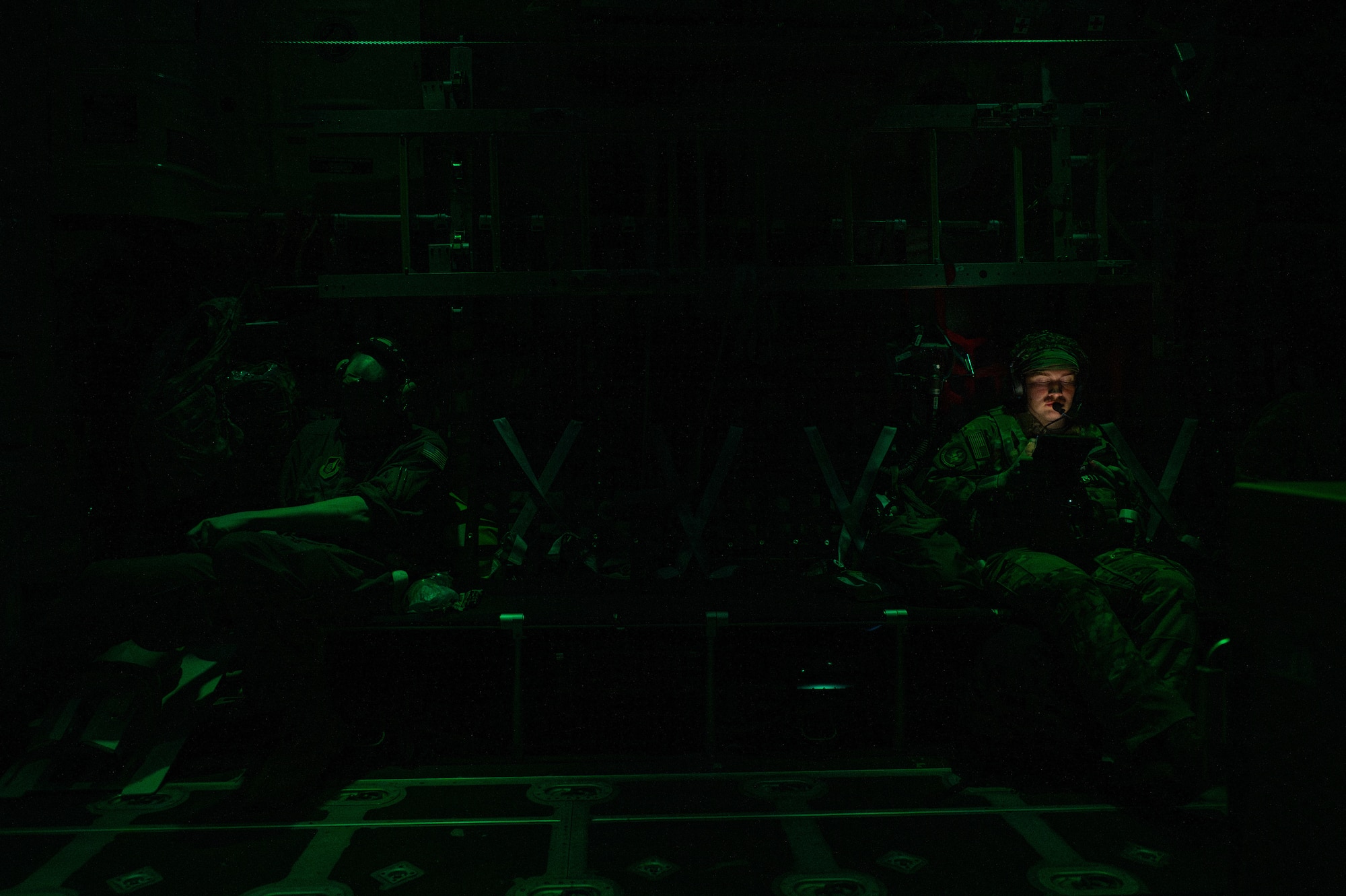 Two military members sit in a MC-130J aircraft hull with the green low light