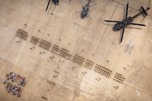 Dozens of soldiers stand in formation in front of three parked helicopters while a seated audience watches as seen from above.