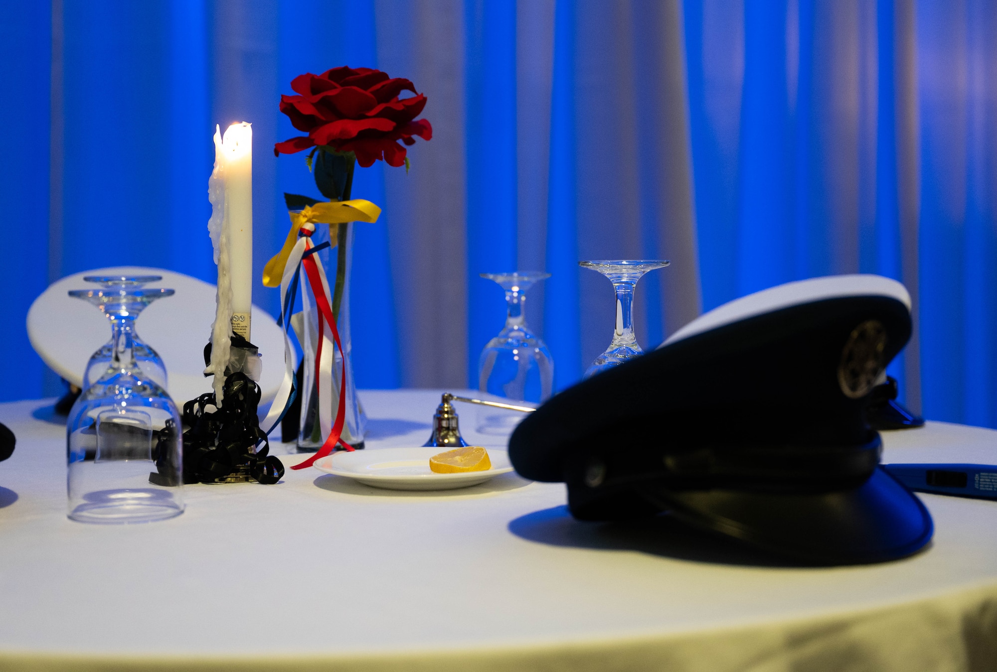 Dress caps of the United States military branches are placed at a table covered in a white cloth a red rose sits in the middle lit by candle light