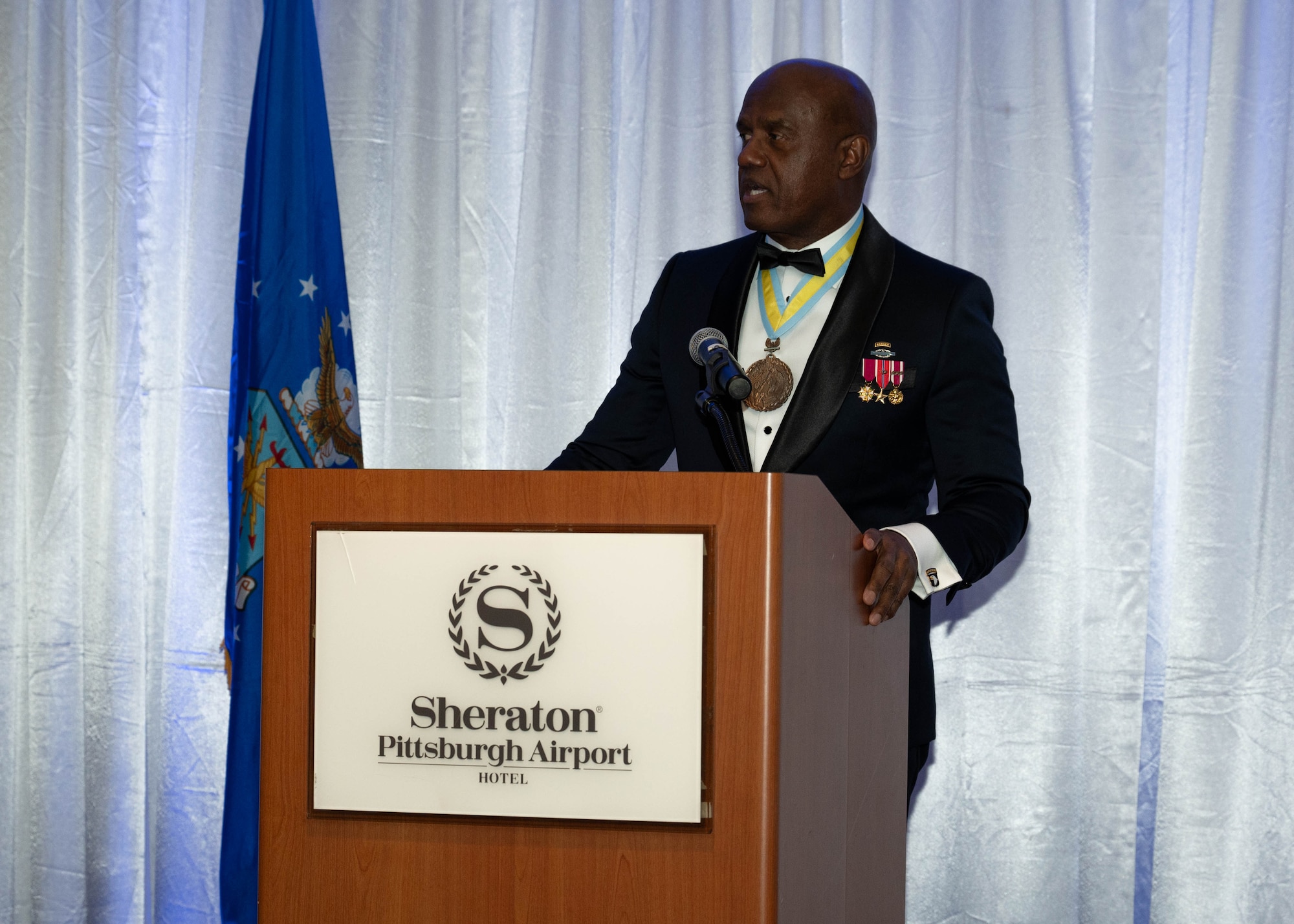 an African American male in a black tuxedo wears military medals and speaks at an awards banquet